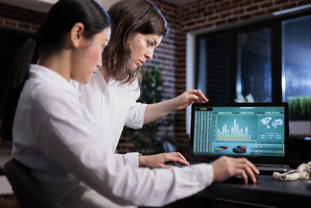 En la fotografía se observa a dos profesionales de una agencia de marketing analizando intensamente datos financieros en la pantalla de un portátil. Sentados frente a un escritorio bien equipado, discuten estrategias de inversión en acciones, quizás centradas en el stock picking, mientras revisan el estado de un proyecto o inicio de empresa. La escena refleja un momento crítico de toma de decisiones, donde el conocimiento avanzado en finanzas y mercados es clave, y la gestión de riesgos es esencial para evitar una potencial bancarrota. El enfoque de los empleados y la complejidad de los gráficos subrayan la seriedad de su tarea en el dinámico entorno del mercado de valores.