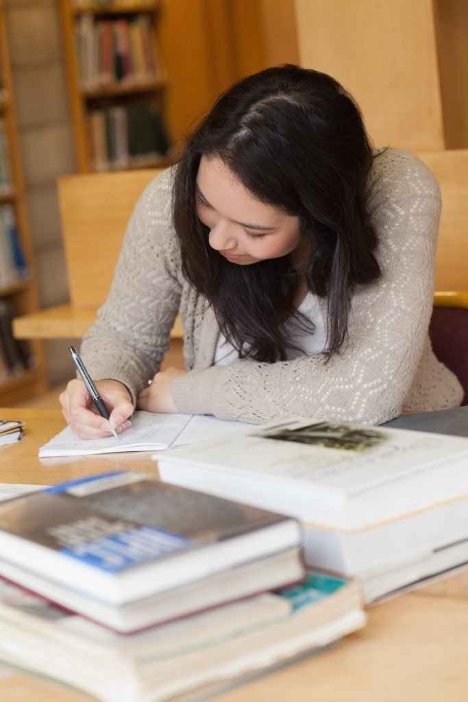 Estudiante aplicada escribiendo notas de Educación Financiera intensamente en su cuaderno, rodeada de libros apilados en una biblioteca.